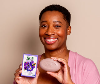 A smiling person in a pink top holds a purple Fresh Violet Moisturizing Bar Soap box with a flower design and matching embossed soap featuring organic shea butter for rich lather, all set against a beige background.