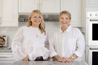 Two women with blonde hair, both wearing white blouses, stand smiling in a bright kitchen with white cabinets and marble countertops. They are leaning casually on the counter, facing the camera.