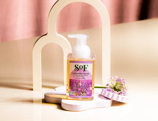 A bottle of Lavender Fields Foaming Hand Soap, paraben and SLS free, rests on a decorative surface beside a purple flower and pink brush, with an abstract cream-colored arch in the background.