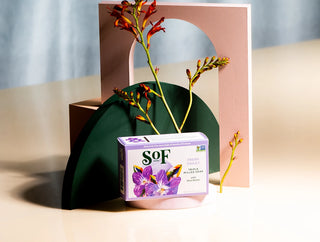A box of Fresh Violet Moisturizing Bar Soap rests on a beige surface, surrounded by abstract geometric shapes and orange flowers, with a soft blue and pink background.