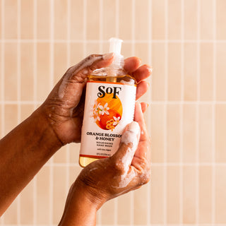 With soapy hands, someone holds Orange Blossom & Honey Nourishing Hand Wash against a beige tiled background.