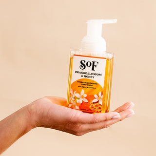 A hand holds a clear pump bottle of Orange Blossom & Honey Hydrating Foaming Hand Wash with aloe vera, orange and white flower graphics on the label, against a beige background. The formula is paraben free and SLS/SLES free.