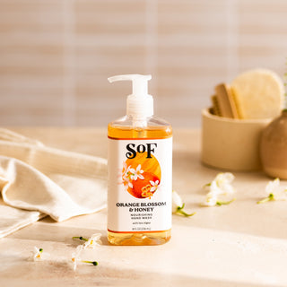 A clear pump bottle of Orange Blossom & Honey Nourishing Hand Wash, free from SLS/SLES, sits on a beige surface with white flowers. In the softly blurred background are a towel and a container holding sponges.