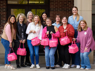 A group of smiling women stand together outside a building, holding bright pink tote bags. Most are dressed casually in jackets and sweaters. They appear happy and are gathered closely for the photo.