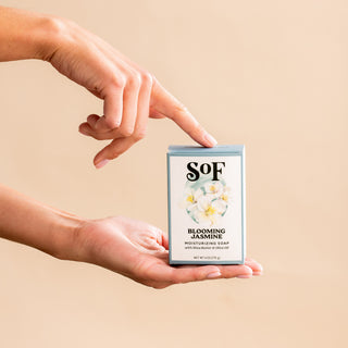 A person holds a box of Blooming Jasmine Moisturizing Bar Soap in one hand and points to it with the other. The box displays white jasmine flowers on a light background.