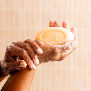 A close-up of two hands holding the light yellow Blooming Jasmine Moisturizing Bar Soap, embossed with “SOF.” The triple-milled soap creates a rich lather and is enriched with organic shea butter. A soft, blurred striped background appears behind.