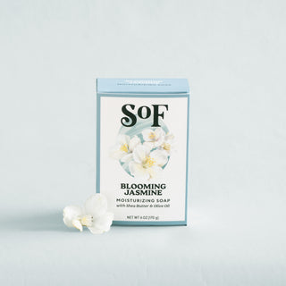 A box of Blooming Jasmine Moisturizing Bar Soap stands upright on a light background, with two white jasmine flowers placed next to it.