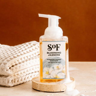 A bottle of Blooming Jasmine Hydrating Foaming Hand Wash rests on a beige stone tray beside neatly stacked white towels, set against a warm brown background.