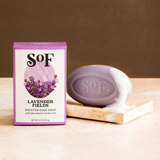 A purple box of Lavender Fields Moisturizing Bar Soap stands beside a lathered lavender bar on a beige stone slab, set against a brown background.