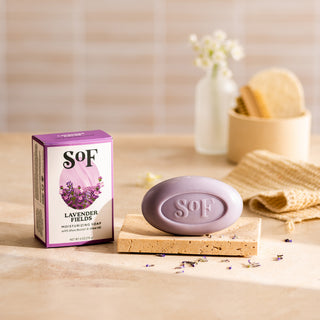 The Lavender Fields Moisturizing Bar Soap, paraben free and colored with natural colorants, rests on a stone tray with dried petals, beside a washcloth and brush; its box is labeled SOF Lavender Fields.