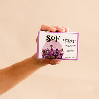 A hand holds a box of Lavender Fields Moisturizing Bar Soap, made with shea butter, olive oil, and natural colorants. The packaging features lavender flowers and purple accents on a neutral background.