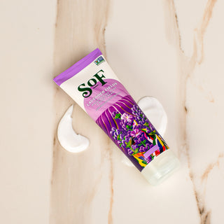 A purple tube of Lavender Fields Hand and Body Cream rests on marble with two dollops of white lotion beside it. The tube displays lavender art, a Non-GMO Project Verified label, and highlights Fair Trade shea butter.