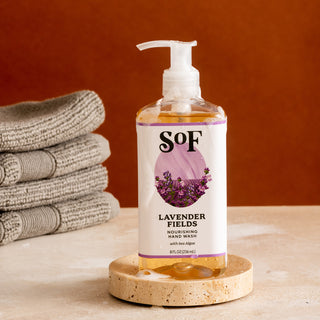 A clear pump bottle of Lavender Fields Nourishing Hand Wash rests on a round stone tray beside folded gray towels on a beige surface against a brown background.