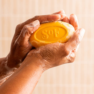 Two soapy hands hold a yellow Lemon Verbena Moisturizing Bar Soap against a light tiled background. This triple-milled soap creates a rich, moisturizing lather, leaving your skin soft and nourished.
