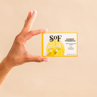 A hand with light pink nails holds a yellow and white box of Lemon Verbena Foaming Hand Soap and Bar Soap against a beige background. The packaging features lemons, verbena flowers, and hints at the soap’s layered scents for daily cleansing.