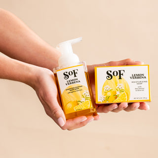 Two hands hold Lemon Verbena Foaming Hand Soap and Bar Soap, both with yellow citrus-themed labels, against a pale beige background—ideal for daily cleansing and enjoying fresh, layered scents.