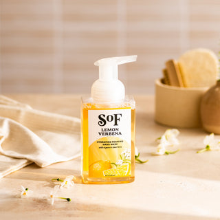 A clear pump bottle of Lemon Verbena Hydrating Foaming Hand Wash, SLS and paraben free, sits on a beige surface with white flowers nearby, plus a towel and sponge container in the background.