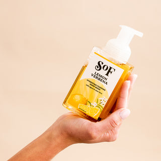 A hand holds a clear bottle of Lemon Verbena Hydrating Foaming Hand Wash with a white pump against a beige background. Lemons and verbena flowers appear on the label, emphasizing its paraben-free and phthalate-free formula.