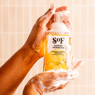 A soapy hand holds a bottle of Lemon Verbena Hydrating Foaming Hand Wash—SLS and paraben free—against a light, tiled background.