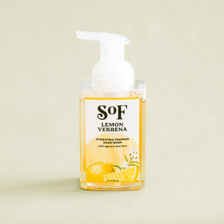A clear bottle of Lemon Verbena Hydrating Foaming Hand Wash with a white pump, featuring lemons and verbena leaves on the label. SLS and paraben free, displayed against a light beige background.