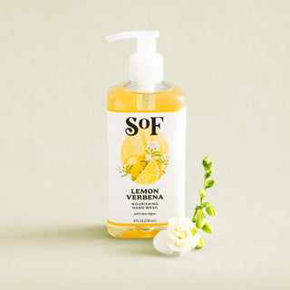 A bottle of Lemon Verbena Nourishing Hand Wash with a pump, featuring a yellow lemon design, is displayed beside a white flower and green bud on a light beige background.