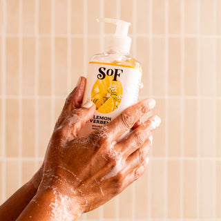 A person’s soapy hands hold a pump bottle of Lemon Verbena Nourishing Hand Wash—free from SLS and parabens—against a tiled bathroom background.