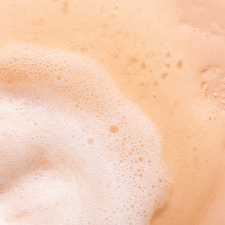 A close-up of the light, creamy foam from the Orange Blossom & Honey Nourishing Hand Wash spreads over a peach-toned surface, dotted with tiny bubbles and subtle background texture.