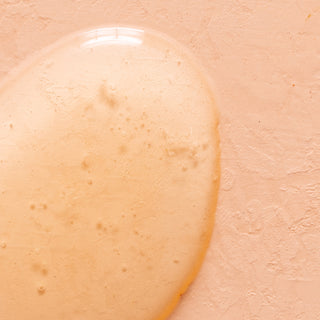 A clear, slightly viscous drop of Lavender Fields Nourishing Hand Wash with small bubbles rests on a textured, peach-colored surface.