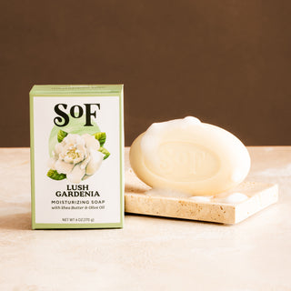A Lush Gardenia Moisturizing Bar Soap with organic shea butter and bubbles sits on a stone soap dish beside its green and white box decorated with a gardenia illustration.