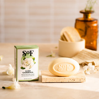 A bar of Lush Gardenia Moisturizing Bar Soap—phthalate and paraben free—rests on a stone tray with its box, white flowers, and a brown glass bottle in the softly lit background for a fresh, elegant look.