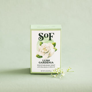 A box of Lush Gardenia Moisturizing Bar Soap, made with shea butter and olive oil, sits on a light green surface. The SLS-free, paraben-free soap features a white gardenia design and a sprig of white flowers beside it.