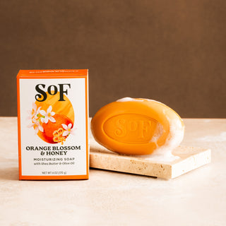 An orange box labeled Orange Blossom & Honey Moisturizing Bar Soap stands beside an orange oval triple-milled soap embossed with "SoF," both on a beige stone surface against a brown background. The soap is slightly lathered.