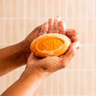 Two hands hold a lathered Orange Blossom & Honey Moisturizing Bar Soap, with the word "Soft" engraved on it, against a light tiled background.
