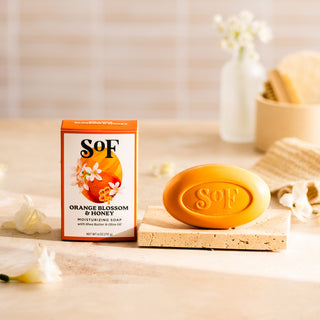 An orange bar from the Orange Blossom & Honey Bundle sits on a stone tray beside its floral-decorated box, with soft lighting, white flowers, and bath accessories in the background.