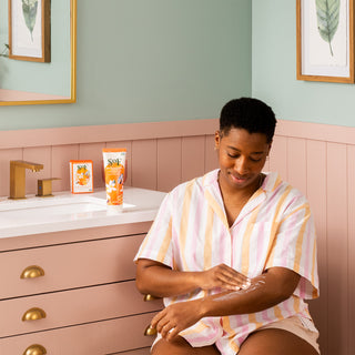 Wearing pastel striped pajamas, a person sits on a bathroom counter applying lotion to their arm, surrounded by Orange Blossom and Honey Bar Soap and Hand & Body Cream atop the white and pink vanity next to the faucet.