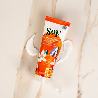 A tube of Orange Blossom & Honey Hand and Body Cream rests on marble, cream swirled beside it. The packaging features oranges, white flowers, and honeycomb accents to highlight its shea and cocoa butter formula.