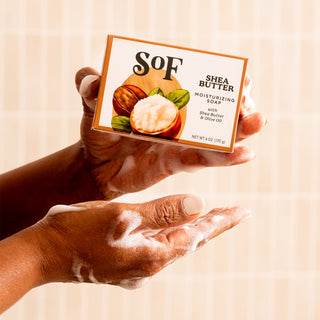 A person with soapy hands holds a bar of Shea Butter Liquid Hand Soap and Bar Soap against beige tiles. The packaging, showing shea nuts and butter, emphasizes its gentle cleansing and nourishing benefits for your skin.