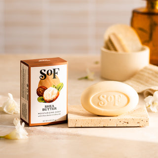 A Shea Butter Moisturizing Bar Soap rests on a stone tray beside its illustrated box, with a jar, towels, and white flowers in the background creating a luxurious display of rich, moisturizing lather.