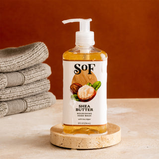 A bottle of Shea Butter Nourishing Hand Wash with a pump is on a round stone tray next to folded gray towels against a brown background.