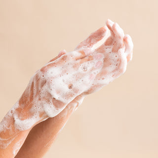 Two hands covered in white soapy foam rub together, using Shea Butter Nourishing Hand Wash, against a plain beige background.