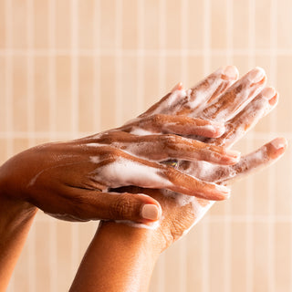 Close-up of two hands interlaced and lathered with Lemon Verbena Nourishing Hand Wash, being washed against a light beige tiled background. The image highlights proper handwashing technique and the product's moisturizing, nourishing lather.