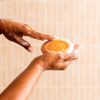 A person’s soapy hands hold a Sweet Almond Moisturizing Bar Soap, embossed with “S&F,” against a light beige tiled background.