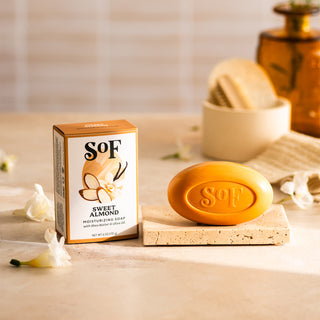 A bar of Sweet Almond Moisturizing Bar Soap rests on a stone tray by its box, with white flower petals nearby on a beige surface. In the background are a rolled towel and an amber glass bottle.