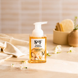 A bottle of Sweet Almond Hydrating Foaming Hand Wash, SLS/SLES free, rests on a beige surface with small white flowers. Behind it, a cloth and a container holding a brush and sponge add to the tranquil setting.