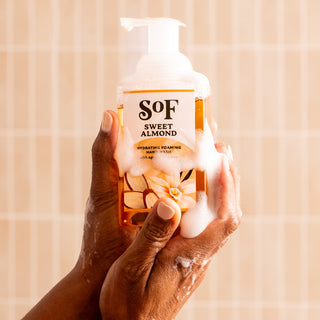 A person holds a clear bottle of Sweet Almond Hydrating Foaming Hand Wash, covered in white suds, against light beige tiles. This gentle hand wash is paraben-free and SLS/SLES-free for healthier skin.