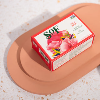 Wild Rose Moisturizing Bar Soap, enriched with organic shea butter, comes in a red and white box with pink flowers and rests on a peach oval dish—ready to deliver a rich, moisturizing lather on any speckled white surface.